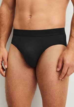 Mann trägt schwarze Unterhose im Brief-Stil mit breitem Bund, gezeigt von der Taille bis zur Mitte des Oberschenkels vor einfarbigem Hintergrund.