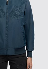 Tummansinihkeä pommikurta, jossa on kukkakirjonta rinnassa, vetoketjukiinnitys sekä resorit hihoissa ja helmassa. Silkkinen kangas.
