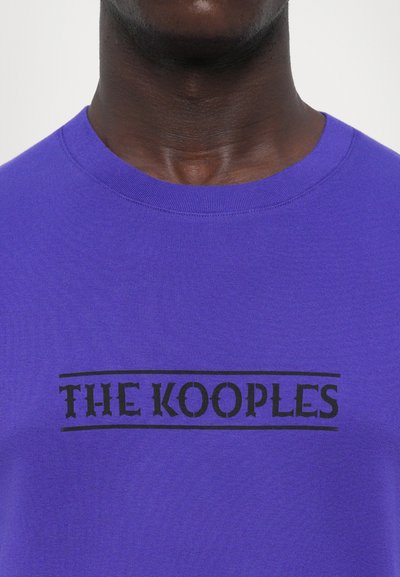 Μωβ βαμβακερό T-shirt με μαύρο ορθογώνιο λογότυπο που γράφει "THE KOOPLES" σε έντονη, στυλιζαρισμένη γραφή. Λείος υφασμάτινος σχεδιασμός και σχέδιο με στρογγυλή λαιμόκοψη.