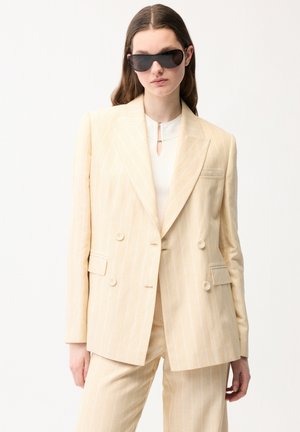 Kvinde iført oversized beige dobbeltradet habit med tynde striber, matchende bukser og store sorte solbriller, stående foran ensfarvet baggrund.