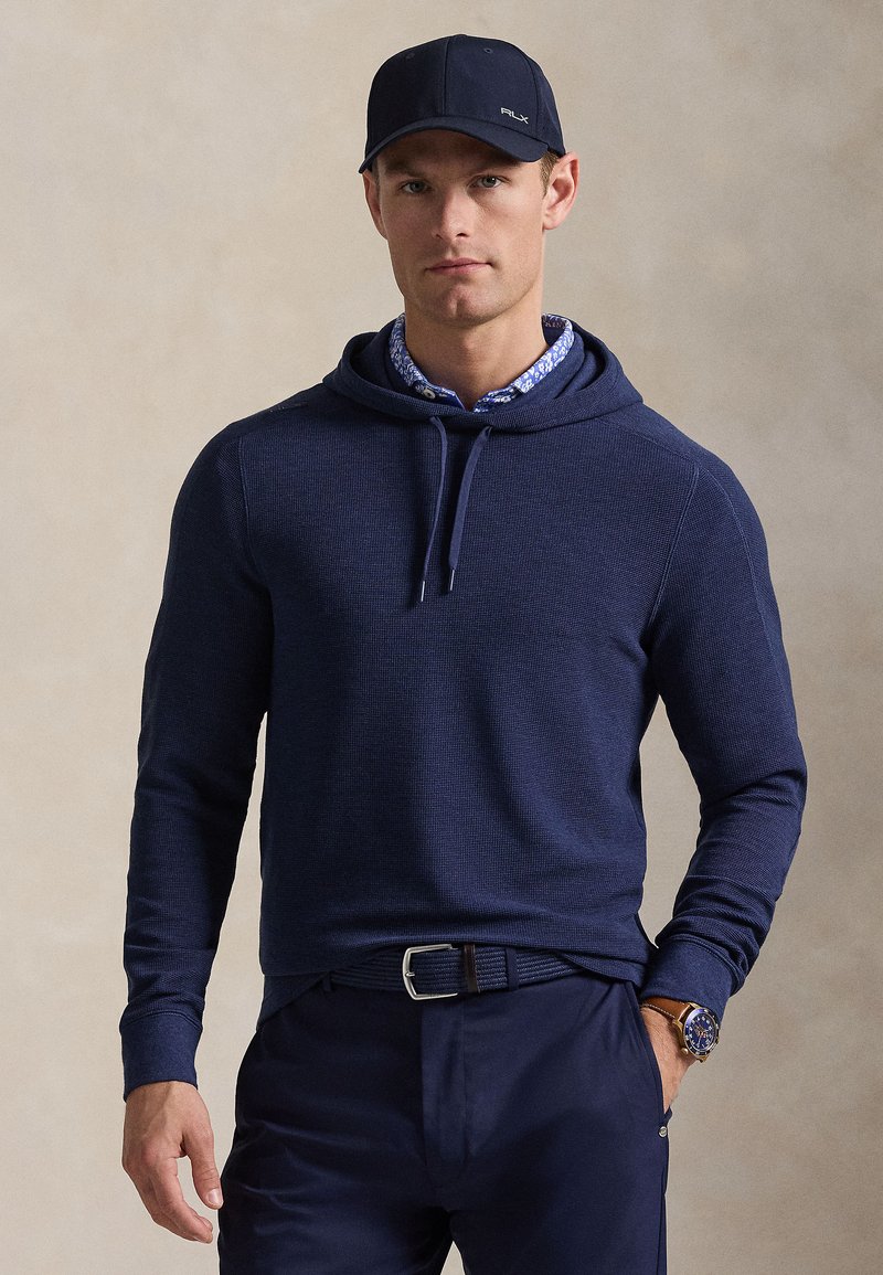 Μπλε σκούρο φούτερ με υφασμένο μοτ ίβο, κουκούλα με κορδόνια και μακριά μανίκια, σε συνδυασμό με μπλε πουκάμισο με γιακά και λουλουδάτο σχέδιο και σκούρο παντελόνι.