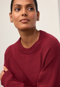 Bordeauxrød sweater med rund halsudskæring, ribbede manchetter og blød tekstur. Bæreres over en neutral baggrund med hænder delvist synlige.