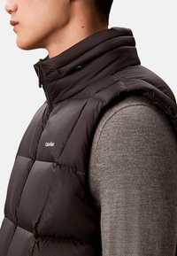 Czarna pikowana kamizelka z wysokim kołnierzem i przeszywanym wzorem, wyposażona w przedni zamek błyskawiczny oraz detal z logo. Noszona na szarym swetrze.