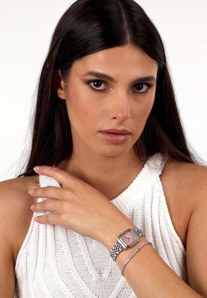 Orologio quadrato con quadrante rosa, cinturino in metallo argento e bracciale con riflessi scintillanti su una maglia bianca testurizzata.