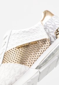 Zapatilla blanca con paneles texturizados y perforados, acentos dorados y logotipo en relieve. Amortiguación transparente en el talón para mayor soporte.