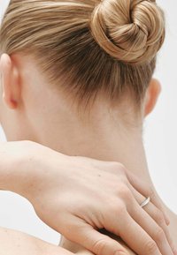 Anillo de plata en una mano descansando sobre el cuello. Cabello peinado en un moño liso y redondo. Piel texturizada con pequeñas pecas; fondo neutro.