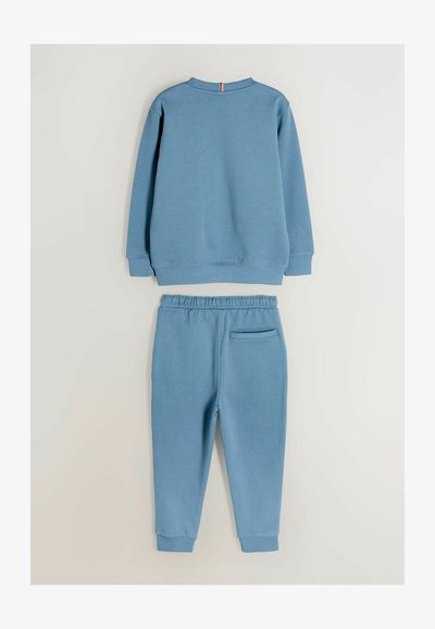 Zila kokvilnas sporta krekls un atbilstošas bikses ar garām piedurknēm, ribotām aprocēm un aizmugurē esošu jostas kabatu. Vienkāršs dizains ar minimāliem akcentiem.