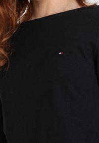 Schwarzes Langarmoberteil aus Baumwolle mit rundem Ausschnitt und einem kleinen Logo mit roten, weißen und blauen Akzenten auf der linken Brust.