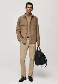 Tan wollen shirt jacket met twee borstzakken over een bijpassende coltrui, gecombineerd met tan broek en zwarte leren schoenen, met een zwarte tas in de hand.