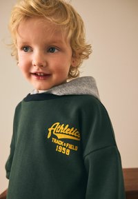 Green sweatshirt with a gray hood, featuring yellow embroidered text "Athletics Track & Field 1998" on the front. Soft cotton material.