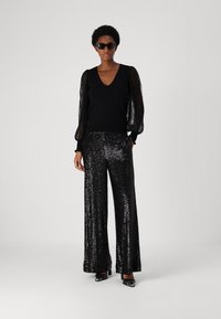 Black long-sleeve top with sheer textured sleeves, paired with high-waisted black sequined wide-leg pants, worn with black heels and sunglasses.