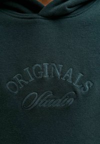 Sudadera con capucha verde oscuro que presenta texto bordado "ORIGINALS" y "Studio", con un acabado suave y texturizado y capucha ajustable con cordón.