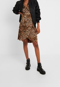 Vestido de estampado de leopardo con escote en V y silueta fluida, combinado con una chaqueta bomber negra y botas negras tipo tobillo con hebillas.