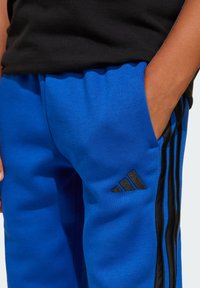Pantalons de sport bleus en tissu doux, avec des bandes latérales noires et un patch logo Adidas noir sur la cuisse gauche.