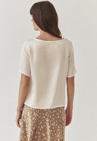 Blusa de manga corta en crema con un amplio escote redondeado y textura suave, combinada con una falda beige que presenta manchas abstractas claras.