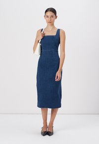 Modrá denimová midi šaty se čtvercovým výstřihem, fitted korzetem a rovnou sukní. Modelka nese bílou kabelku a má na sobě černé podpatky.