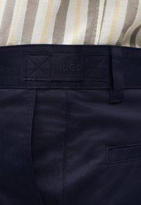 Σκούρο μπλε στενά παντελόνια με θηλιές για ζώνη, μια πίσω τσέπη με πλαινό άνοιγμα και ένα ραμμένο έμπλαστρο με την ένδειξη «HUGO» πάνω από τη μέση.