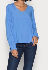 Kvinna iklädd en långärmad himmelsblå blus med volangmanchetter, tillsammans med mörkblå åtsittande jeans.