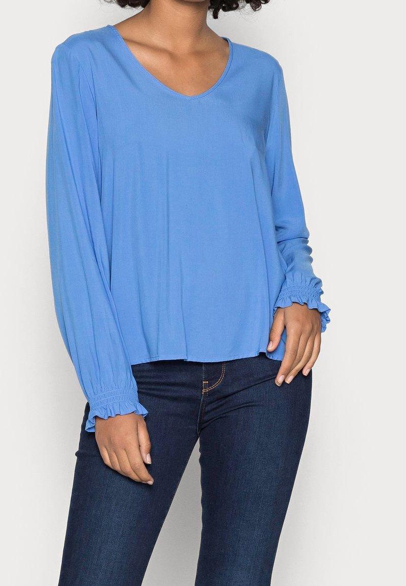 Kvinna iklädd en långärmad himmelsblå blus med volangmanchetter, tillsammans med mörkblå åtsittande jeans.