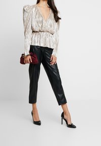 Metallic, pleated beige top with a deep V-neck and elastic waist, paired with black leather tapered pants and a burgundy clutch bag. Black heels.