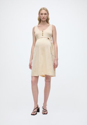 Έγκυος γυναίκα φοράει μπεζ αμάνικο φόρεμα μήκους μέχρι το γόνατο με λεπτομέρειες από κουμπιά και μαύρα ψηλοτάκουνα σανδάλια, στέκεται μπροστά σε ένα απλό λευκό φόντο.