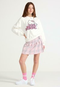 Felpa bianca con grafica rosa di Hello Kitty, abbinata a una gonna a pieghe a quadri rosa. Sneakers bianche e calzini a strisce rosa completano il look.