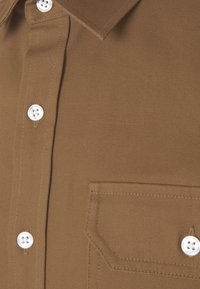 Chemise en tissu texturé marron avec des boutons blancs et une poche poitrine avec des coutures visibles.