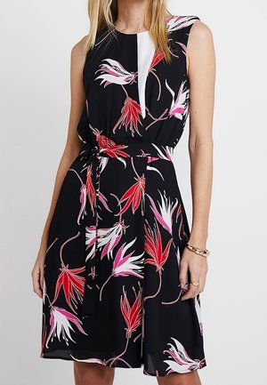 Czarna sukienka bez rękawów w czerwono-biało-różowy kwiatowy wzór, z talią podkreśloną wiązanym paskiem, noszona przez stojącą kobietę na jednolitym tle.