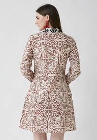 Pitkähihainen mekko vaaleassa värissä, jossa on punainen abstrakti kuvio. Fitted vyötärö ja suora siluetti, jossa on kontrastinen musta kaulus.