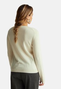 Suéter de punto crema claro con cuello redondo y puños acanalados, mostrado desde atrás, combinado con pantalones gris oscuro. Textura suave.