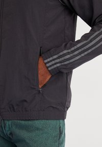 Black lightweight jacket with a zippered pocket, featuring elastic cuffs and three white stripes along the sleeves. Textured fabric.