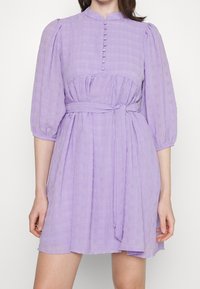 Lila Kleid mit hohem Kragen, puffartigen Ärmeln, Knopfleiste und tailliertem Gürtel. Der strukturierte Stoff zeigt ein Karomuster.