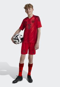 Punainen Bayern Münchenin jalkapallovarustus, jossa on V-mallinen paita, mustat yksityiskohdat, shortsit, korkeat sukat, mustat nappulit ja musta-valkoinen jalkapallo.