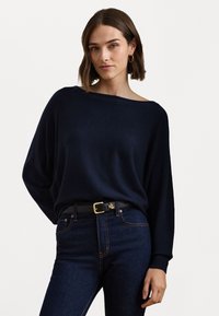 Tengerészkék, laza szabású pulóver, puha anyagból, széles nyakkivágással és bordás mandzsettával. Sötét denim farmernadrággal és fekete övvel párosítva.