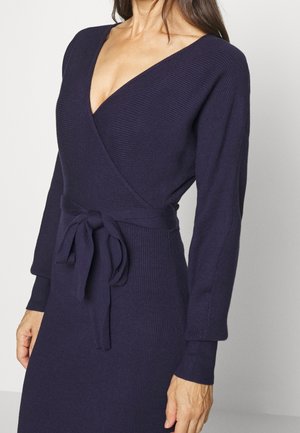 Robe en tricot côtelé bleu marine foncé avec un décolleté en V profond, des manches longues et une taille nouée. Présente une silhouette ajustée et un tissu texturé.