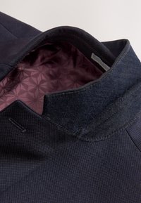 Tumesinine jakk tekstuurse väliskanga ja pehme, mustrilise burgundi voodriga. Omadusteks on tahke krae ja kontrastsed õmblused.