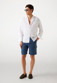 Chemise blanche en lin à manches longues, shorts cargo bleus, chaussures à enfiler marron et lunettes de soleil. Look décontracté avec une poche sur la poitrine et des poches cargo.