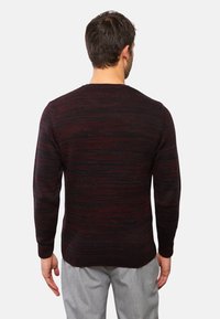 Uomo che indossa un maglione a righe nero e bordeaux scuro e pantaloni grigi chiari, in piedi rivolto di spalle contro uno sfondo neutro.