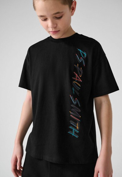 Garçon portant un T-shirt noir surdimensionné avec le texte coloré "PAUL SMITH" imprimé verticalement sur le devant, regardant vers le bas.