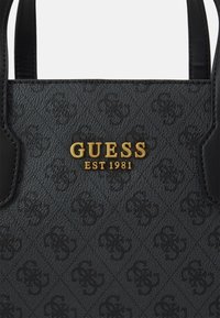 Černá texturovaná kabelka se zlatým logem s nápisem "GUESS EST 1981", která se vyznačuje podrobným embosováním a kontrastními koženými akcenty.