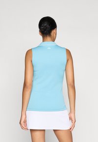 Camisola polo sem mangas de cor azul claro, com textura canelada e acabamento branco na gola. Um logótipo aparece perto do pescoço. Usada com uma saia branca.