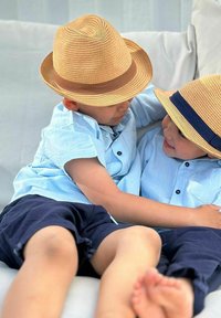 Kaksi lasta istuu lähekkäin, päällään vaaleansiniset lyhythihaiset paidat, tummat shortsit ja olkihatut, joissa on ruskeita ja tummansiniä nauhoja.