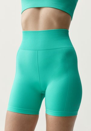 Close-up af en person iført turkis ribstrikkede træningsshorts med høj talje og en matchende sports-bh mod en ensfarvet baggrund.