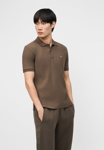 Άνδρας με καφέ πόλο πουκάμισο και παντελόνι στο ίδιο χρώμα, με τα χέρια στις τσέπες, κοιτάζοντας προς τα αριστερά, στέκεται μπροστά σε απλό ανοιχτό φόντο.