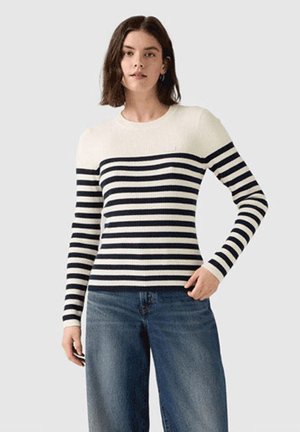 Mujer que lleva un suéter blanco de manga larga con rayas horizontales negras y vaqueros azules, de pie con una mano en el bolsillo.