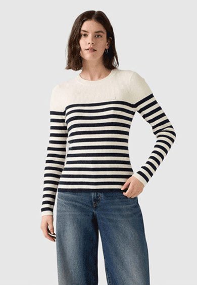 Woman wearing a long-sleeve white sweater with black horizontal stripes and blue jeans, standing with one hand in her pocket.