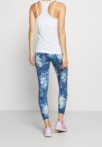 Canotta atletica bianca con spalline sottili, abbinata a leggings blu tie-dye e sneakers rosa chiaro, vista da dietro.