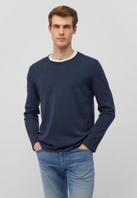 Dunkelblaues Langarmshirt mit Rundhalsausschnitt, aus glattem Stoff. Kombiniert mit hellblauen Jeans, vor einem neutralen Hintergrund stehend.