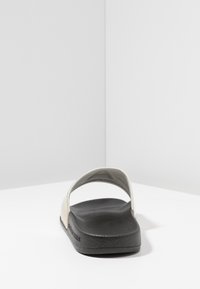 Sandały typu slide z białą cholewką i czarną teksturowaną podeszwą, z otwartym tyłem i szerokim paskiem, co ułatwia zakładanie.