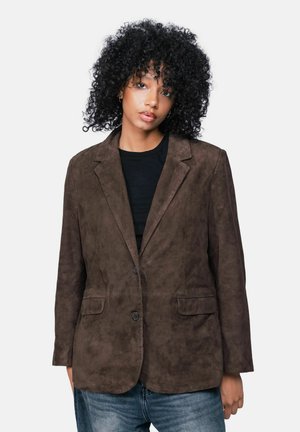 Femme aux cheveux noirs bouclés portant un blazer en daim brun foncé par-dessus un top noir et un jean bleu sur un fond uni.